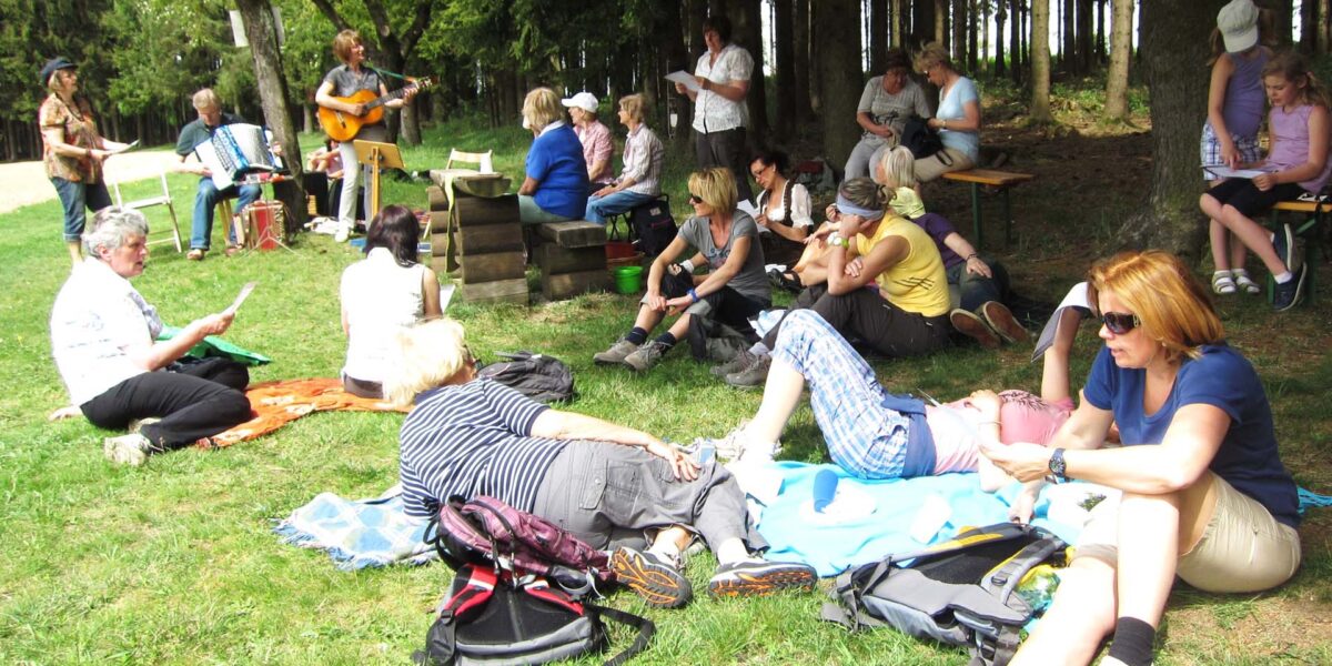 Eine Gruppe von Menschen entspannt sich auf dem Rasen und auf Bänken in einem Park, während im Hintergrund Musiker Akkordeon und Gitarre spielen.