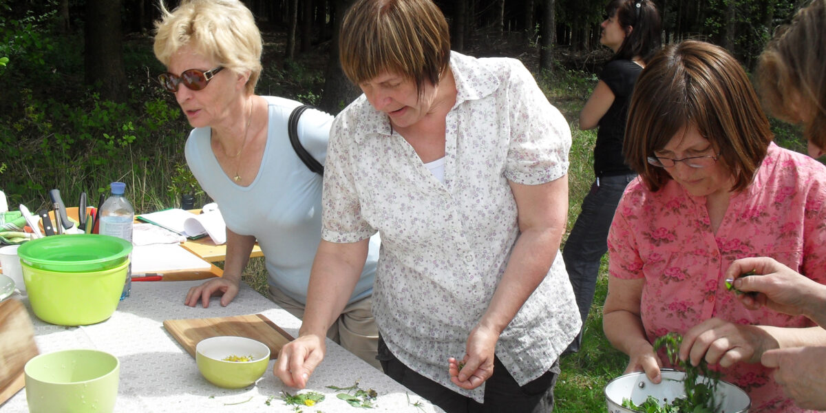 Drei Frauen bereiten Kräuter an einem Tisch im Freien mit Schüsseln und Schneidebrettern zu, während andere in einem Waldstück zusehen.