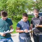Drei junge Männer stehen an einem sonnigen Tag im Freien, schauen auf Papiere und halten Stifte in der Hand, im Hintergrund sind Bäume und Gebäude zu sehen.