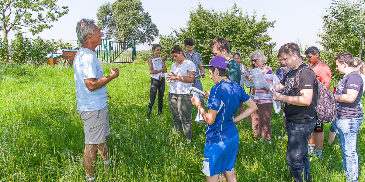 Eine Gruppe von Menschen steht im Freien auf einer Wiese und hört einem Mann zu, der Papiere in der Hand hält. Im Hintergrund sind Bäume und ein Zaun zu sehen.