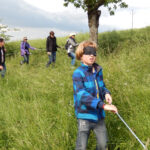 Eine Gruppe von Menschen läuft mit verbundenen Augen durch hohes Gras und hält sich an einem Seil fest, das sie führt.