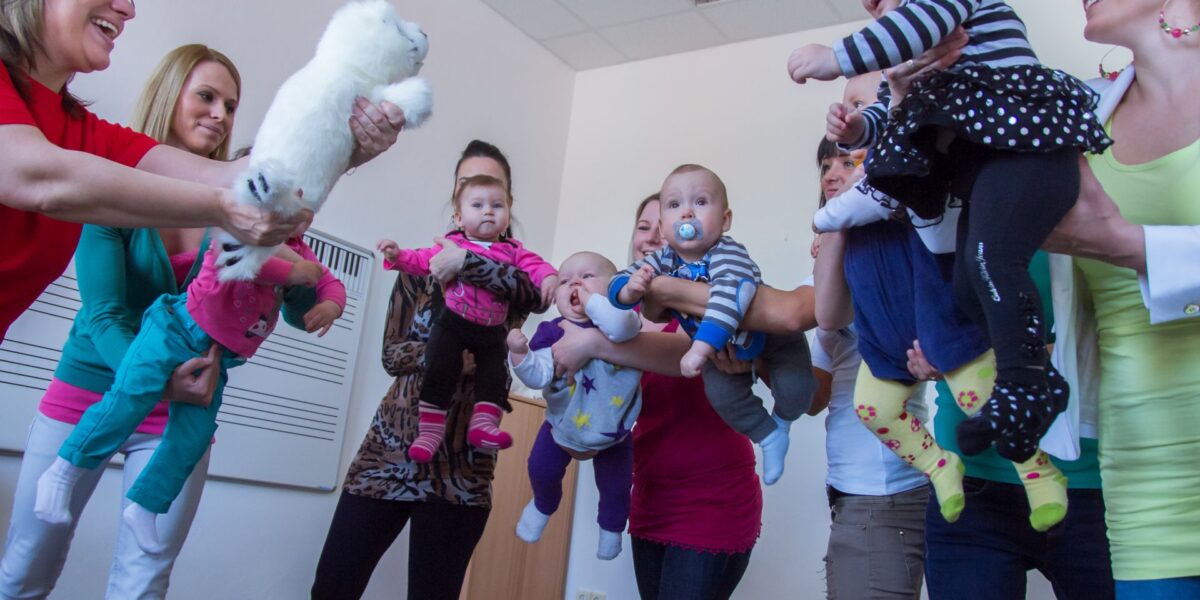 Eine Gruppe Erwachsener hält Babys in einem Innenraum im Kreis, wobei ein Erwachsener ein weißes Stofftier in Richtung der Babys hält.