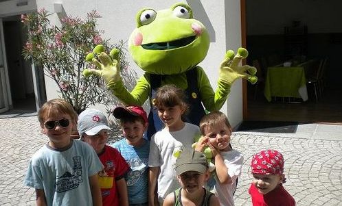 Eine Gruppe von sechs kleinen Kindern posiert draußen mit einer Person in einem großen grünen Froschkostüm, die hinter ihnen steht, ihre Hände hebt und lächelt.