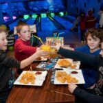 Vier Jungen sitzen an einem Tisch in einer Bowlingbahn, stoßen mit Getränken an und essen Pommes und Chicken Nuggets.