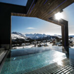 Infinity-Pool im Freien mit Blick auf die Berge, moderne architektonische Struktur über dem Wasser, strahlender Sonnenschein, schneebedeckte Landschaft im Hintergrund.