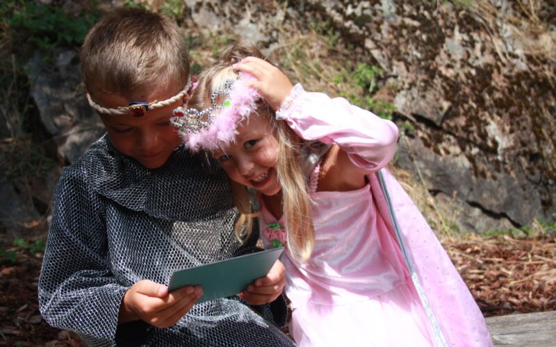 Ein Junge und ein Mädchen in Kostümen, der eine als Ritter, der eine Karte liest, und die andere als Prinzessin, die lächelt und ihre Krone zurechtrückt, sitzen im Freien zusammen.