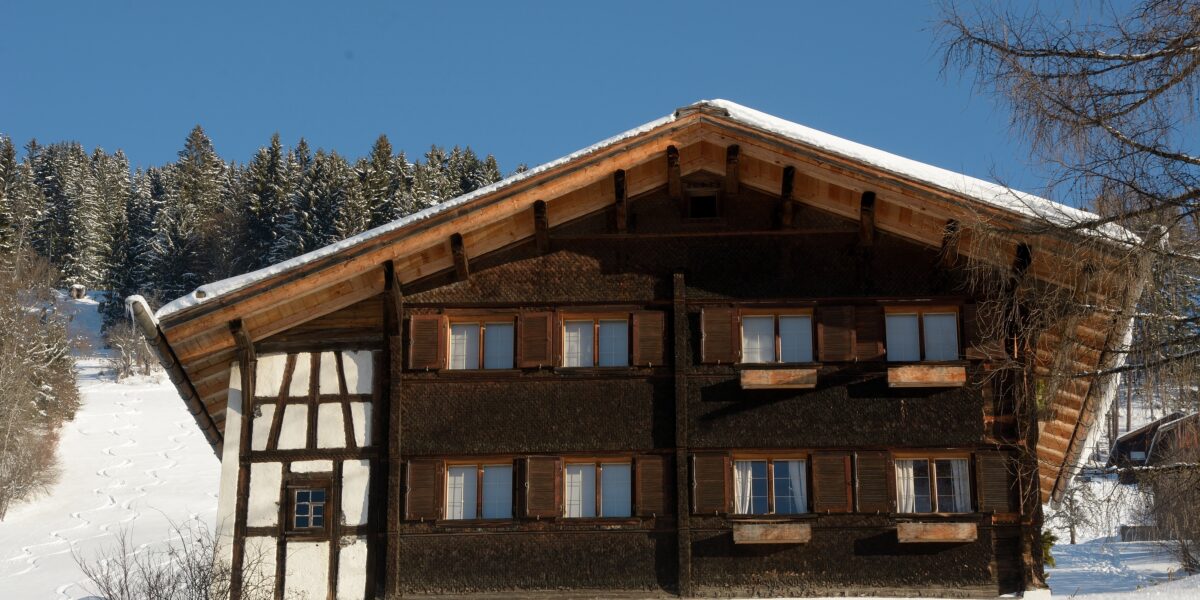 Ein traditionelles Holzchalet mit geschlossenen Fensterläden steht in einer verschneiten Landschaft, umrahmt von Bäumen unter einem klaren blauen Himmel.