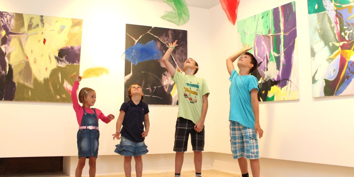 Vier Kinder stehen in einer Kunstgalerie und werfen bunte Tücher in die Luft, während hinter ihnen abstrakte Gemälde an den Wänden hängen.