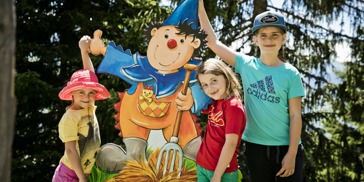 Drei Kinder stehen an einem sonnigen Tag im Freien neben einem bunten Cartoon-Ausschnitt eines Gnoms, der eine Mistgabel hält, umgeben von Bäumen.