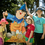 Drei Kinder stehen an einem sonnigen Tag im Freien neben einem bunten Cartoon-Ausschnitt eines Gnoms, der eine Mistgabel hält, umgeben von Bäumen.