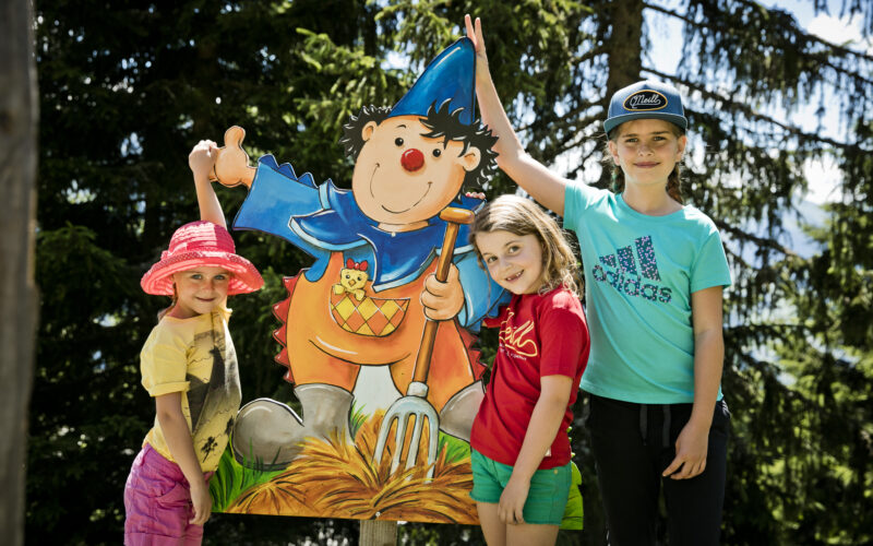 Drei Kinder stehen an einem sonnigen Tag im Freien neben einem bunten Cartoon-Ausschnitt eines Gnoms, der eine Mistgabel hält, umgeben von Bäumen.