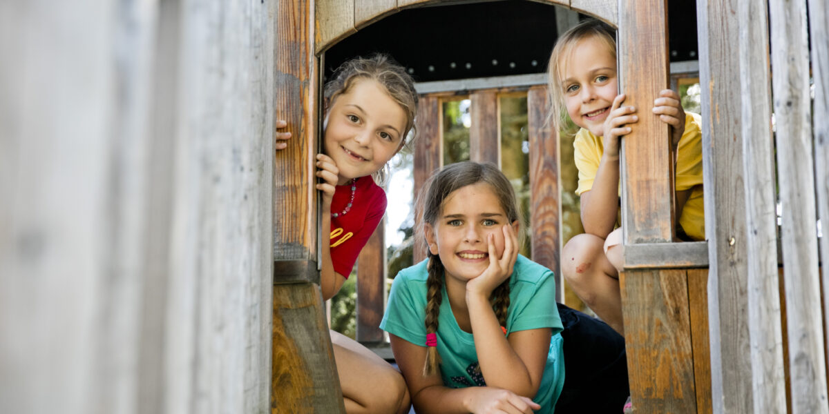 Drei Kinder im Inneren eines hölzernen Spielgerüsts, die lächelnd durch eine offene Tür hinausschauen.