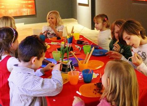 Eine Gruppe von Kindern sitzt um einen Tisch mit bunten Tellern und Tassen, isst und unterhält sich in einem hell erleuchteten Raum.