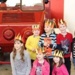 Sieben Kinder mit Papierkronen posieren vor einem alten roten Feuerwehrauto und lächeln für das Foto.