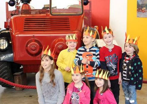 Sieben Kinder mit Papierkronen posieren vor einem alten roten Feuerwehrauto und lächeln für das Foto.