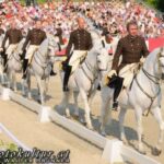 Eine Gruppe von Reitern in braunen Uniformen paradiert auf weißen Pferden in einer Freiluftarena vor einem sitzenden Publikum.
