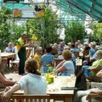 Eine Gruppe von Menschen sitzt an Holztischen in einem Gewächshaus und hört einer Person in grünem Overall zu, die einen Vortrag inmitten von Pflanzen hält.