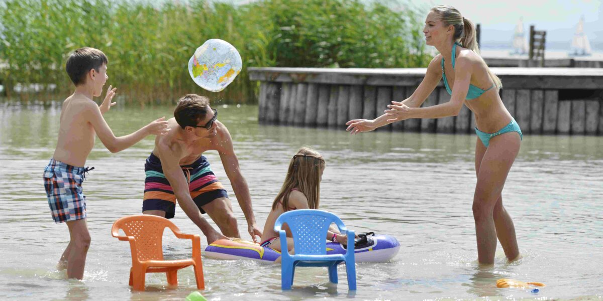 Eine Familie mit zwei Kindern spielt an einem sonnigen Tag mit aufblasbarem Spielzeug und Plastikstühlen im flachen Wasser eines Strandes.