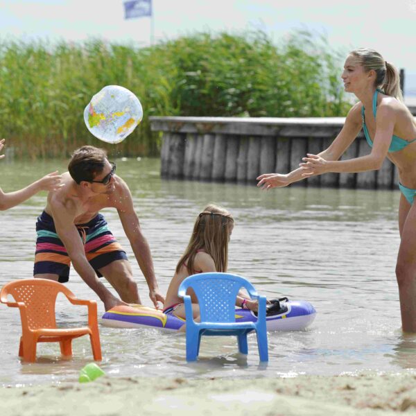 Eine Familie mit zwei Kindern spielt an einem sonnigen Tag mit aufblasbarem Spielzeug und Plastikstühlen im flachen Wasser eines Strandes.