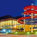 In einem nachts hell erleuchteten Gebäude befindet sich eine große Wasserrutsche im Freien mit roten und blauen Röhren, die sich um das Gebäude winden.
