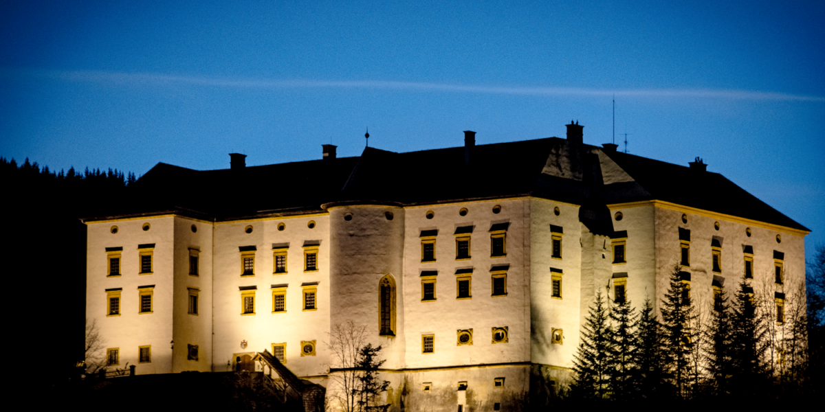 Großes beleuchtetes Schloss mit mehreren Fenstern und Türmchen, umgeben von Bäumen, fotografiert in der Abenddämmerung vor einem tiefblauen Himmel.