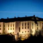 Großes beleuchtetes Schloss mit mehreren Fenstern und Türmchen, umgeben von Bäumen, fotografiert in der Abenddämmerung vor einem tiefblauen Himmel.