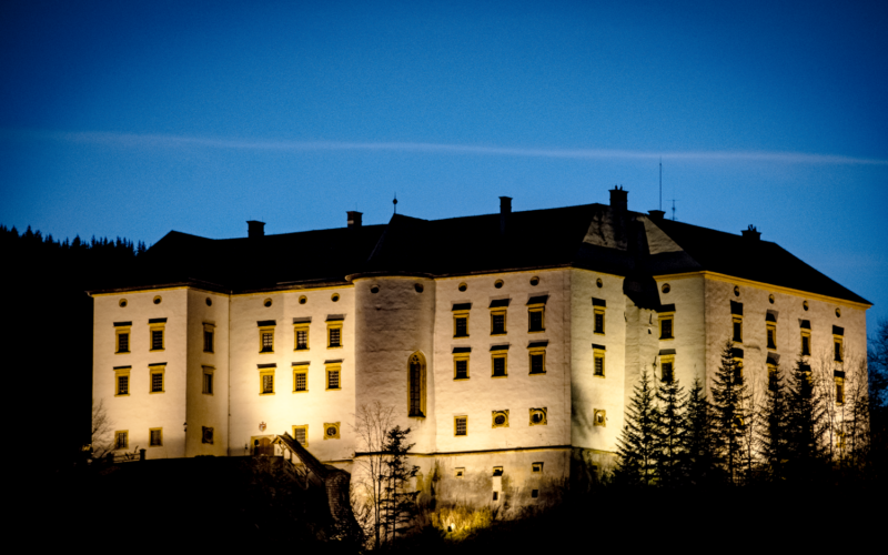 Großes beleuchtetes Schloss mit mehreren Fenstern und Türmchen, umgeben von Bäumen, fotografiert in der Abenddämmerung vor einem tiefblauen Himmel.