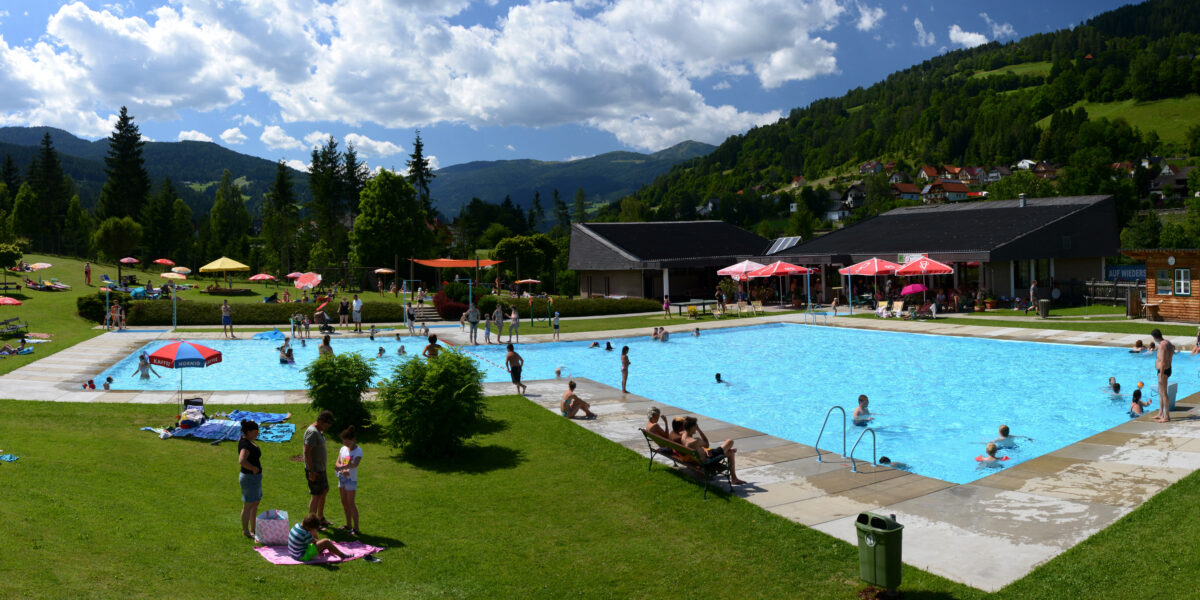 Außenpool mit schwimmenden und entspannenden Menschen; Rasenfläche mit Sonnenschirmen und Liegestühlen; Gebäude und grüne Hügel im Hintergrund unter einem teilweise bewölkten Himmel.