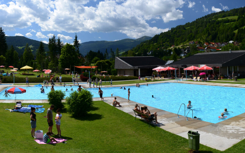 Außenpool mit schwimmenden und entspannenden Menschen; Rasenfläche mit Sonnenschirmen und Liegestühlen; Gebäude und grüne Hügel im Hintergrund unter einem teilweise bewölkten Himmel.