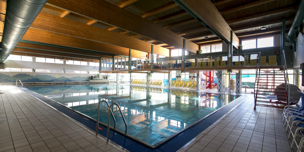 Hallenbad mit klarem Wasser, gefliestem Deck, Metallleitern und gelben Sitzreihen an der Seite, unter einer Holzdecke mit großen Fenstern.