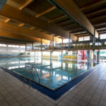 Hallenbad mit klarem Wasser, gefliestem Deck, Metallleitern und gelben Sitzreihen an der Seite, unter einer Holzdecke mit großen Fenstern.