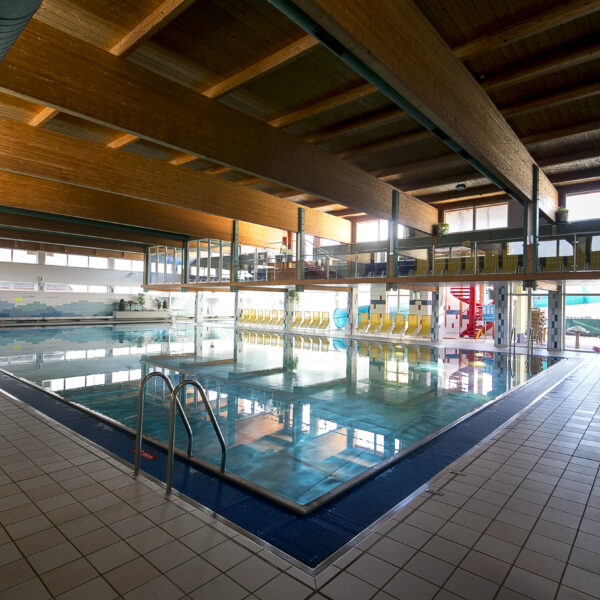 Hallenbad mit klarem Wasser, gefliestem Deck, Metallleitern und gelben Sitzreihen an der Seite, unter einer Holzdecke mit großen Fenstern.