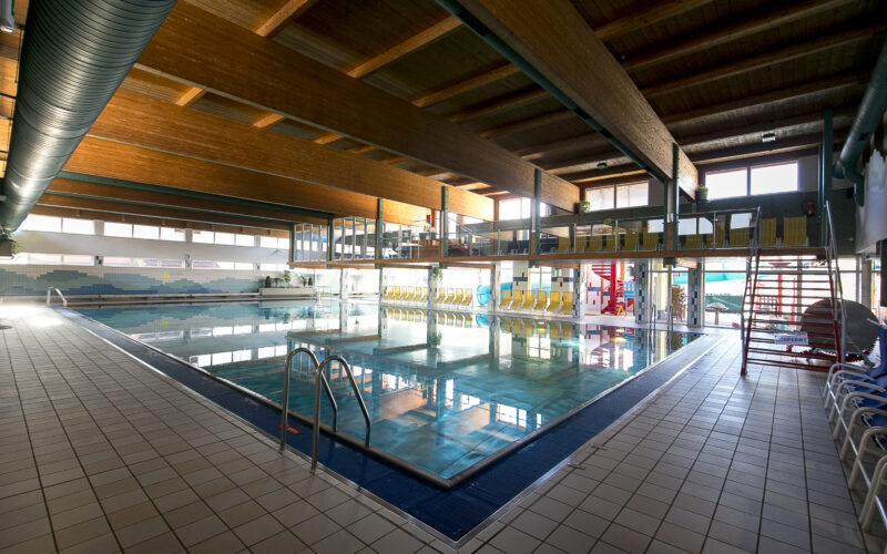 Hallenbad mit klarem Wasser, gefliestem Deck, Metallleitern und gelben Sitzreihen an der Seite, unter einer Holzdecke mit großen Fenstern.