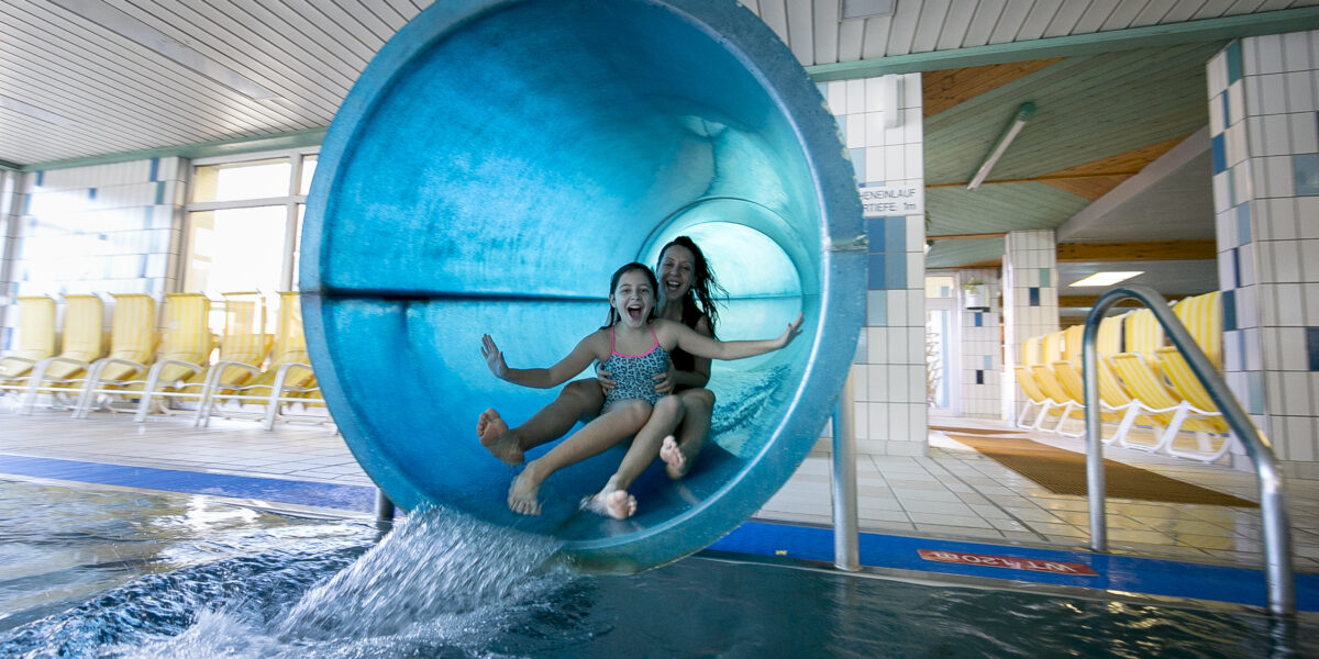 Zwei Kinder, die mit erhobenen Armen lächelnd aus einer blauen Indoor-Wasserrutsche in ein Schwimmbecken steigen, in einer hell erleuchteten Wasseranlage.