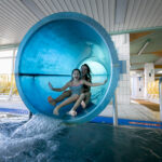 Zwei Kinder, die mit erhobenen Armen lächelnd aus einer blauen Indoor-Wasserrutsche in ein Schwimmbecken steigen, in einer hell erleuchteten Wasseranlage.