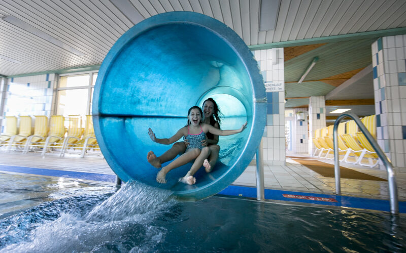 Zwei Kinder, die mit erhobenen Armen lächelnd aus einer blauen Indoor-Wasserrutsche in ein Schwimmbecken steigen, in einer hell erleuchteten Wasseranlage.