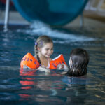 Zwei junge Mädchen schwimmen in einem Pool; ein Mädchen trägt orangefarbene Schwimmflügel und lächelt die andere an, während sie im Wasser in der Nähe einer Rutsche spielen.