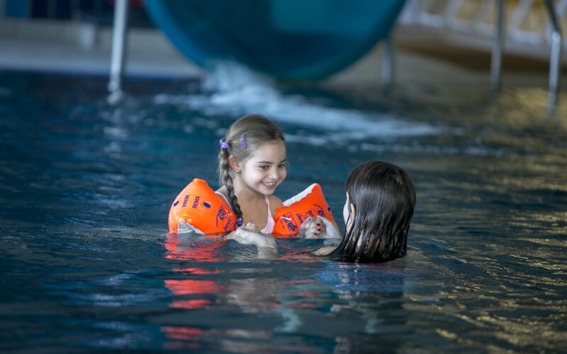 Zwei junge Mädchen schwimmen in einem Pool; ein Mädchen trägt orangefarbene Schwimmflügel und lächelt die andere an, während sie im Wasser in der Nähe einer Rutsche spielen.