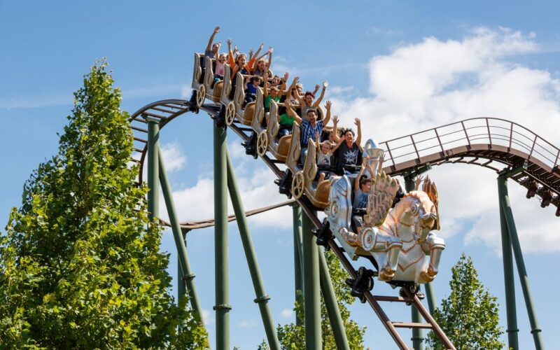 Eine Gruppe von Menschen mit erhobenen Armen fährt auf einer Achterbahn in Form von Pferden eine Strecke im Freien hinunter, umgeben von grünen Bäumen und einem blauen Himmel.