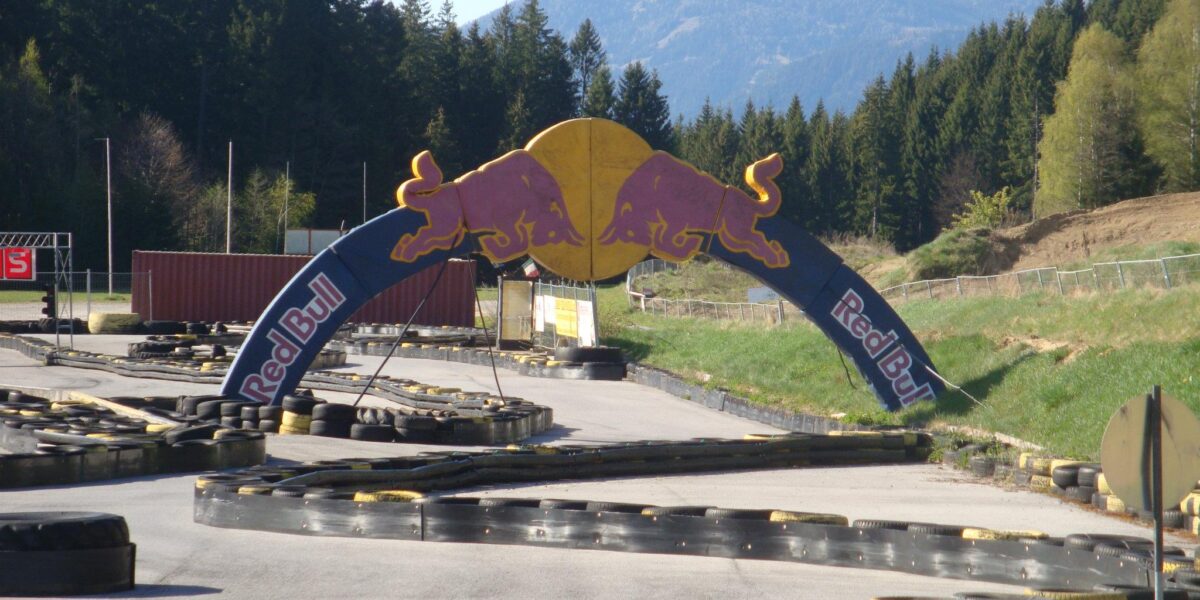 Go-Kart-Bahn im Freien mit einem Red Bull-Bogen im Vordergrund, umgeben von Reifen, Bäumen und Bergen im Hintergrund.