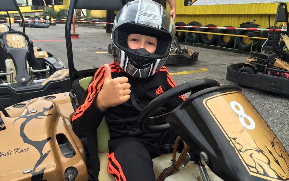 Ein Kind, das einen Helm und einen schwarzen Trainingsanzug mit orangefarbenen Streifen trägt, sitzt in einem Gokart und zeigt den Daumen nach oben. Im Hintergrund sind andere Gokarts und ein gelbes Banner zu sehen.