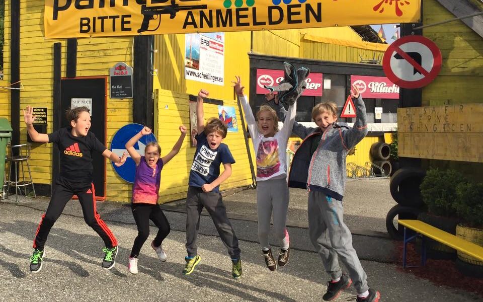 Fünf Kinder springen vor einem gelben Gebäude mit einem Schild, auf dem steht: "Paintball-Areal! Bitte anmelden." Das Wetter scheint sonnig zu sein.