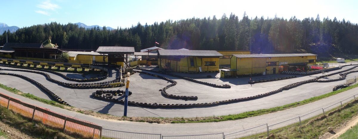 Ein Panoramablick auf eine Go-Kart-Bahn im Freien mit gewundenen Bahnen, umgeben von Bäumen und Bergen und mehreren gelben Gebäuden in der Mitte.