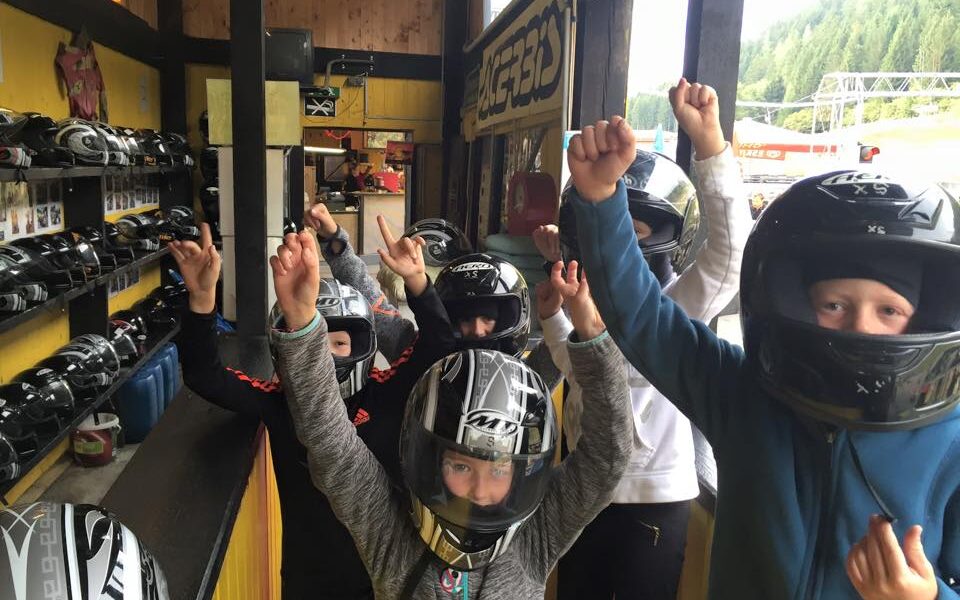 Eine Gruppe von Kindern, die Helme tragen, hebt ihre Hände in einer Go-Kart-Anlage, während im Hintergrund Helme in Regalen ausgestellt sind.