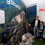 Ein Mann erklärt drei Kindern in einem Museum eine Ausstellung über Erdrutsche. An den Wänden hängen große Fotos von einem felsigen Hang und einem Wald.
