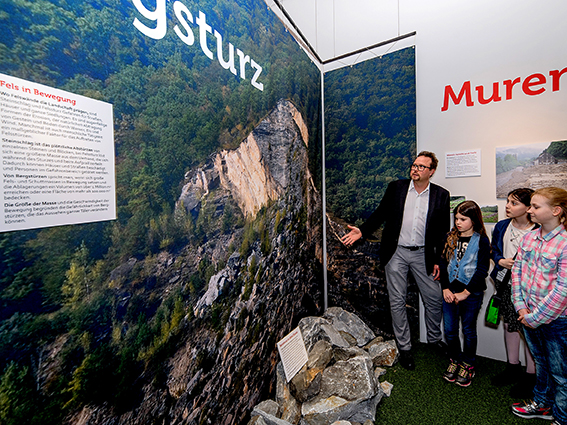 Ein Mann erklärt drei Kindern in einem Museum eine Ausstellung über Erdrutsche. An den Wänden hängen große Fotos von einem felsigen Hang und einem Wald.