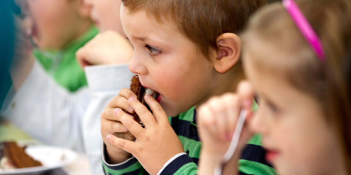 Ein kleiner Junge in einem grün gestreiften Hemd isst ein Stück Brot oder Kuchen und sitzt mit anderen Kindern an einem Tisch.
