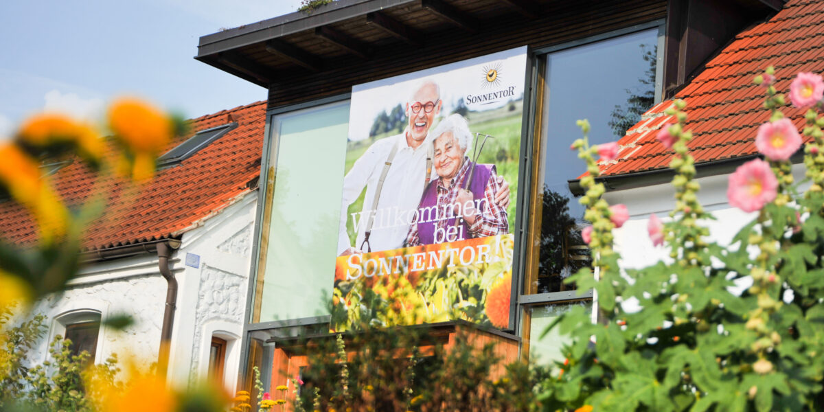 Ein Gebäude mit rotem Ziegeldach trägt ein großes Schild mit einem älteren Ehepaar und der Aufschrift "Willkommen bei Sonnentor", umgeben von Blumen und Grünpflanzen.