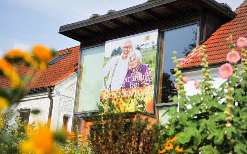 Ein Gebäude mit rotem Ziegeldach trägt ein großes Schild mit einem älteren Ehepaar und der Aufschrift "Willkommen bei Sonnentor", umgeben von Blumen und Grünpflanzen.