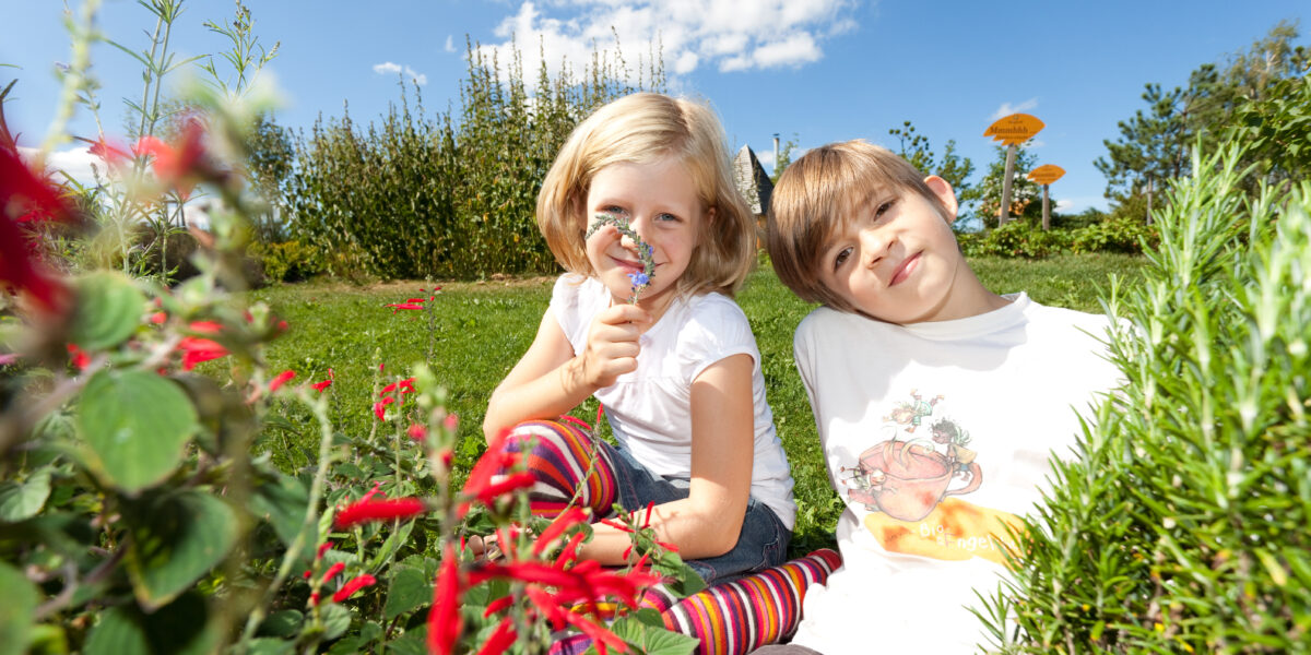 Zwei Kinder sitzen an einem sonnigen Tag im Gras eines Gartens; eines hält sich eine Lupe vors Gesicht, beide sind von roten Blumen und Grün umgeben.